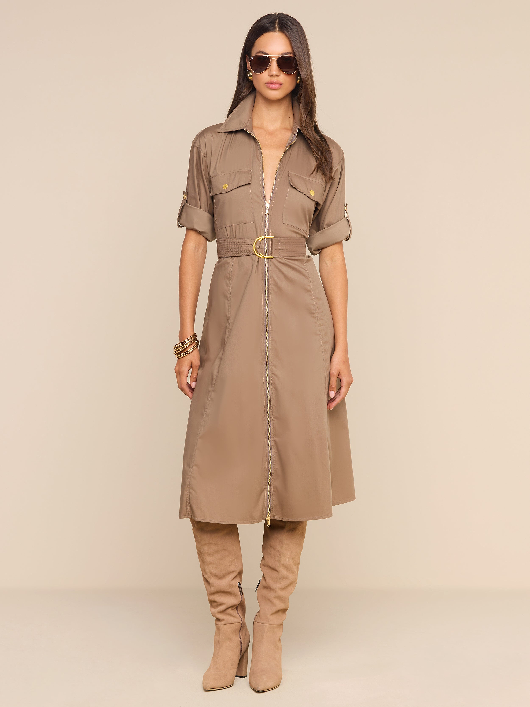 Une femme porte la robe-chemise « Callen » — une robe-chemise en popeline de couleur beige clair, en mélange de coton, avec une ceinture à la taille et des manches retroussées —, quelle a assortie à des bottes beiges claires montant jusquaux genoux. Elle se tient debout devant un fond beige, ses longs cheveux raides sont lâchés, et elle porte des bracelets et des lunettes de soleil.