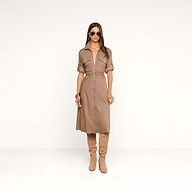 Une femme se tient debout devant un fond beige, vêtue de la robe-chemise « Callen » : une pièce en popeline de coton mélangé couleur beige clair, aux manches retroussées, dotée de boutons dorés et dune fermeture éclair apparente sur le devant. Elle lassocie à des bottes en daim beige clair montant jusquaux genoux, à des lunettes de soleil et à des bracelets dorés.