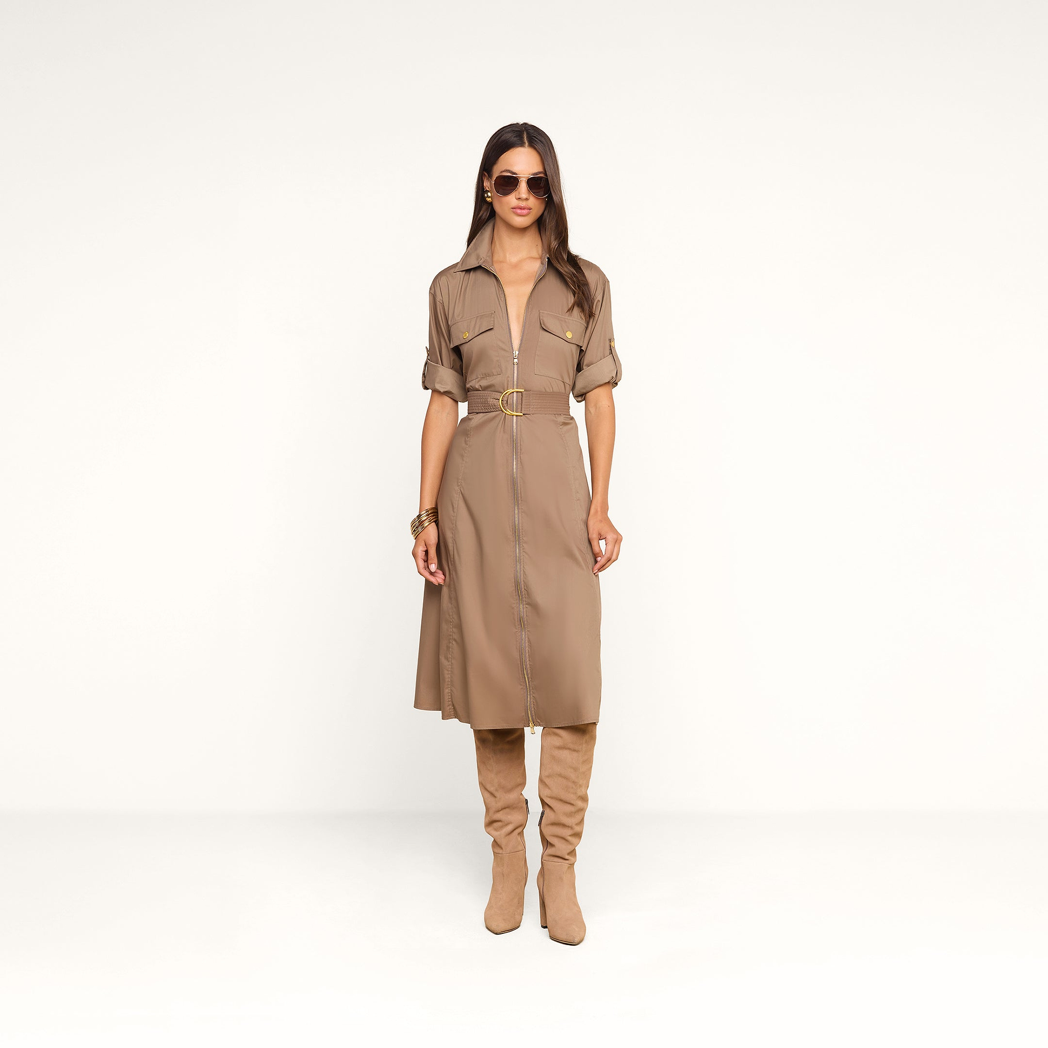Une femme se tient debout devant un fond beige, vêtue de la robe-chemise « Callen » : une pièce en popeline de coton mélangé couleur beige clair, aux manches retroussées, dotée de boutons dorés et dune fermeture éclair apparente sur le devant. Elle lassocie à des bottes en daim beige clair montant jusquaux genoux, à des lunettes de soleil et à des bracelets dorés.
