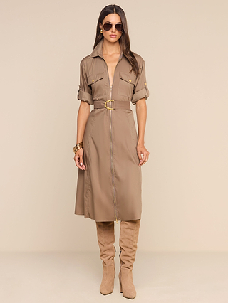 A woman stands against a beige background wearing the Callen Shirt Dress—a tan cotton-blend poplin piece with rolled sleeves, gold buttons, and an exposed front zip. She pairs it with knee-high tan suede boots, sunglasses, and gold bangles.