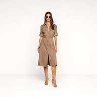 Une femme se tient debout devant un fond beige uni, vêtue de la robe-chemise Callen : un modèle ceinturé, arrivant aux genoux, en popeline de coton mélangé couleur beige clair, doté dune fermeture éclair apparente sur le devant, de manches courtes et de poches. Elle complète sa tenue avec des lunettes de soleil, des bracelets et des sandales à talons hauts.