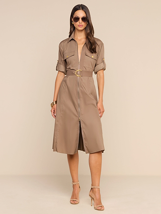 A woman stands against a plain beige background wearing the Callen Shirt Dress—a belted, knee-length design in tan cotton-blend poplin, featuring an exposed front zip, short sleeves, and pockets. She completes her look with sunglasses, bracelets, and high-heeled sandals.