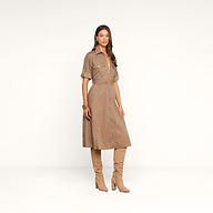 Vêtue de la robe-chemise Callen — une robe-chemise beige clair à col et manches courtes, en popeline de coton mélangé, dotée dune fermeture éclair apparente sur le devant et dune ceinture —, la femme se tient debout devant un fond beige uni, chaussée de hautes bottes en daim beige et parée daccessoires dorés.