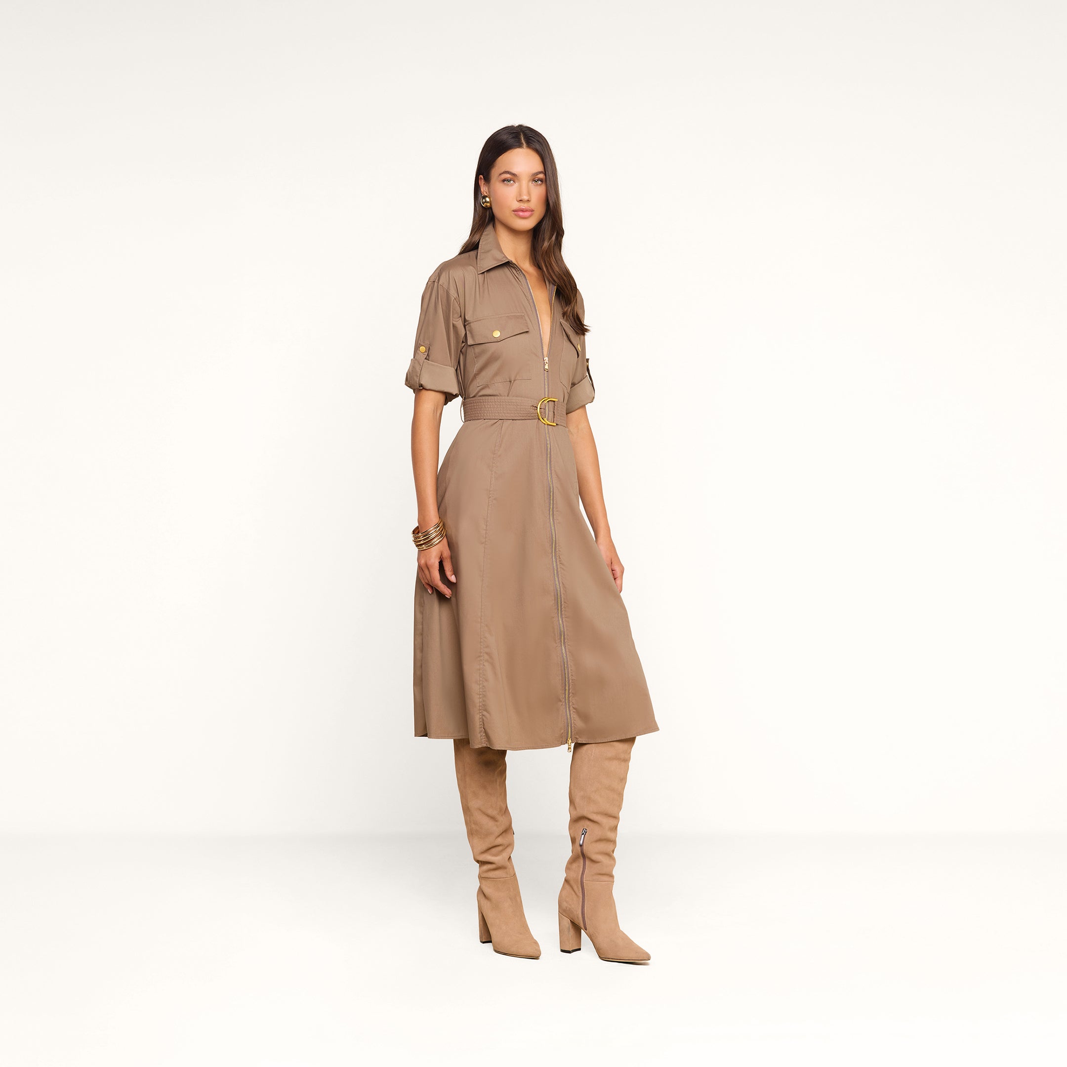 Vêtue de la robe-chemise Callen — une robe-chemise beige clair à col et manches courtes, en popeline de coton mélangé, dotée dune fermeture éclair apparente sur le devant et dune ceinture —, la femme se tient debout devant un fond beige uni, chaussée de hautes bottes en daim beige et parée daccessoires dorés.