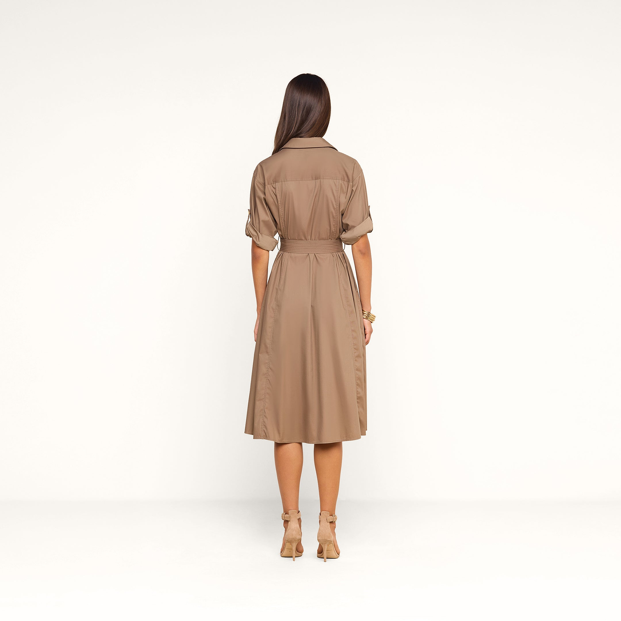Une femme aux longs cheveux châtains se tient dos à la caméra devant un fond beige. Elle porte la robe-chemise « Callen » — une robe-chemise couleur beige clair, dotée d’une ceinture et d’un col, confectionnée en popeline de coton mélangé et aux manches retroussées —, assortie à des escarpins beiges.