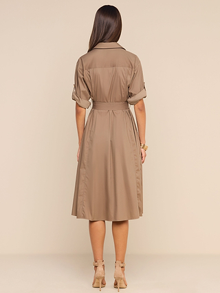 A woman with long brown hair stands with her back to the camera against a beige background, wearing the Callen Shirt Dress—a tan, belted, collared shirt dress made from cotton-blend poplin with rolled-up sleeves—paired with beige heels.