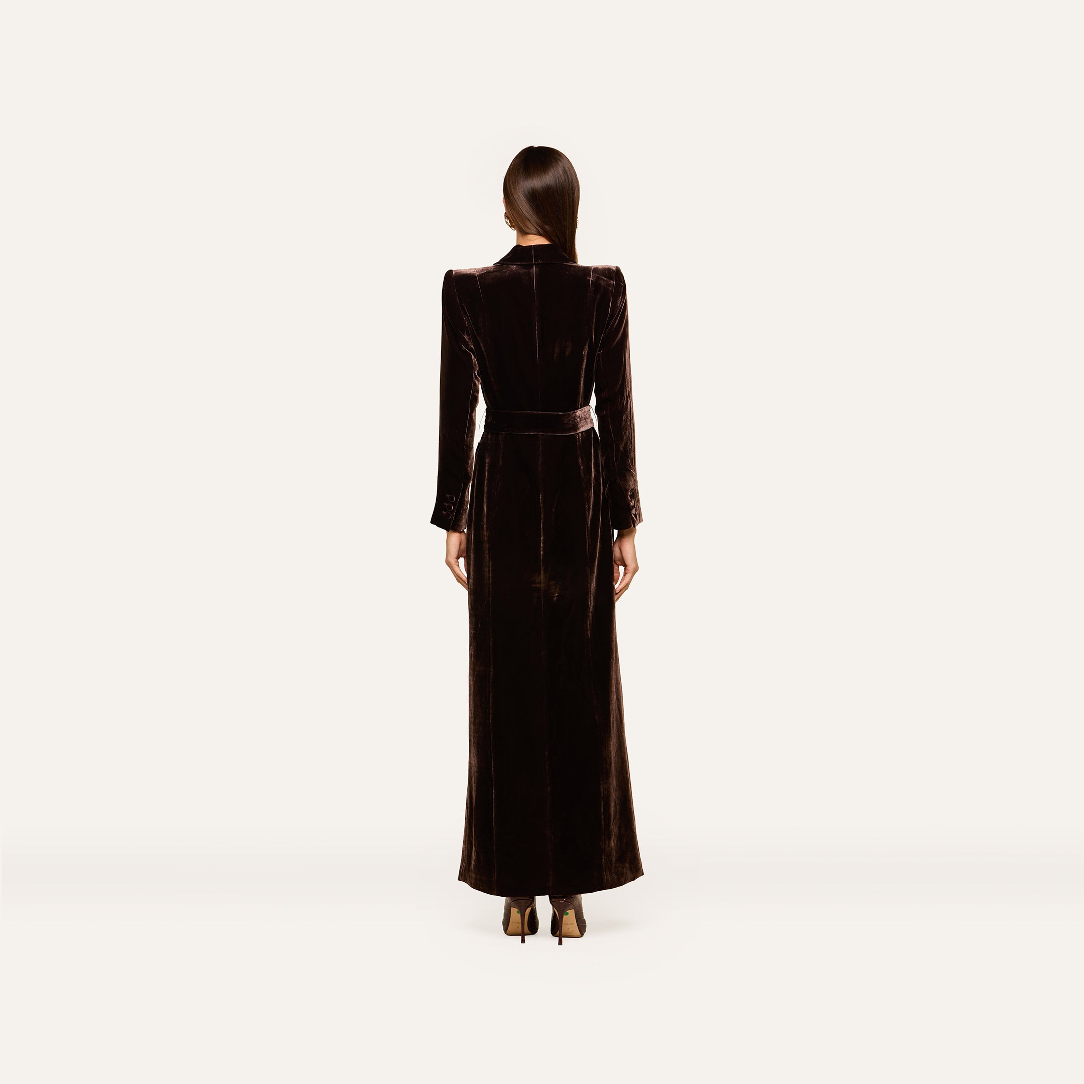 A woman stands facing away, dressed in the Raini Velvet Maxi Dress featuring a shawl collar, long sleeves, and a belted waist. She pairs it with dark heels and has her straight hair tied back. The plain, light-colored background highlights the elegance of Fall 2026 fashion.