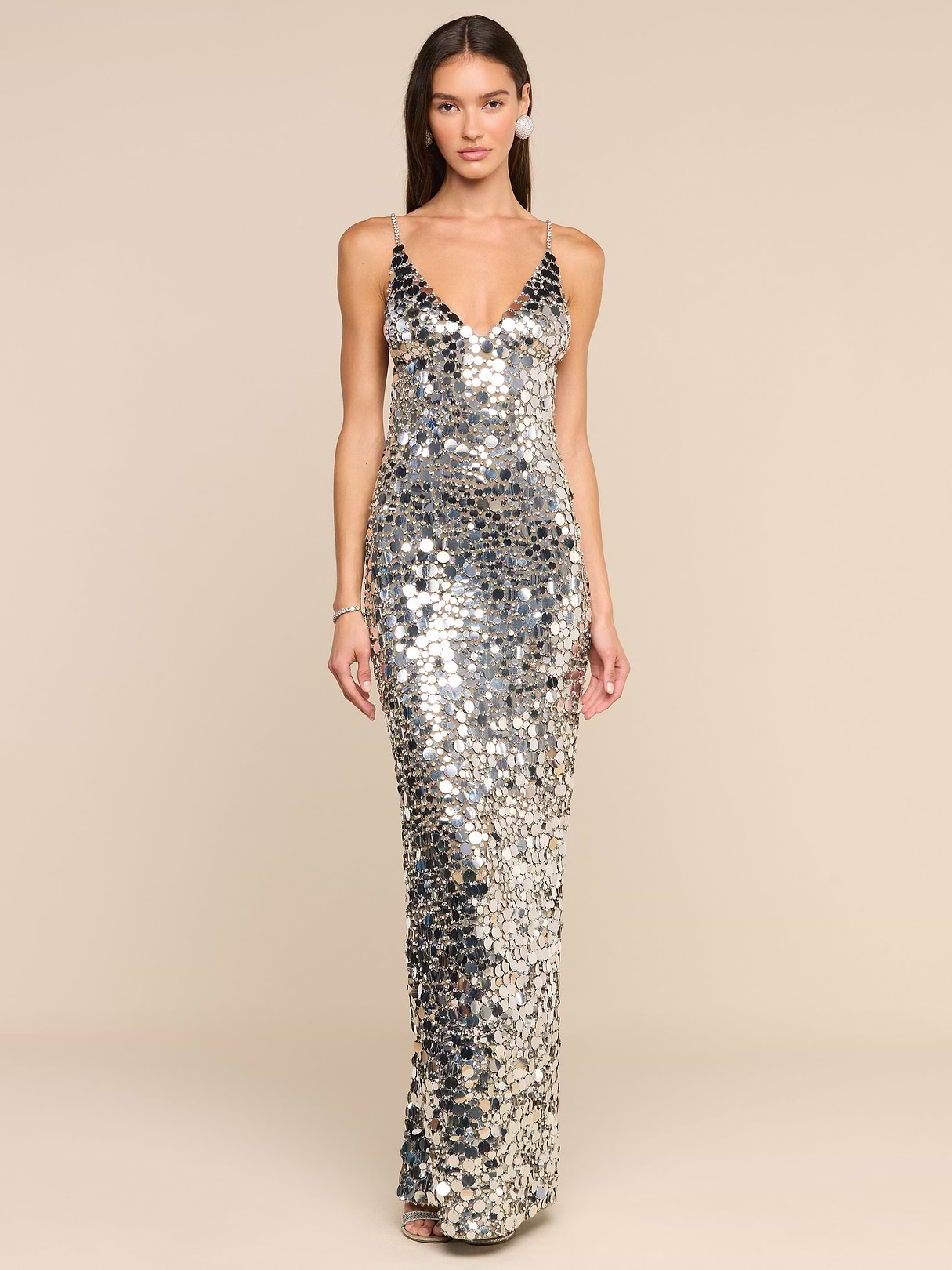 A woman stands facing forward against a plain white background, wearing the Caelith Sequin Gown—a floor-length, sleeveless evening dress in silver with thin straps and shimmering sequins.
