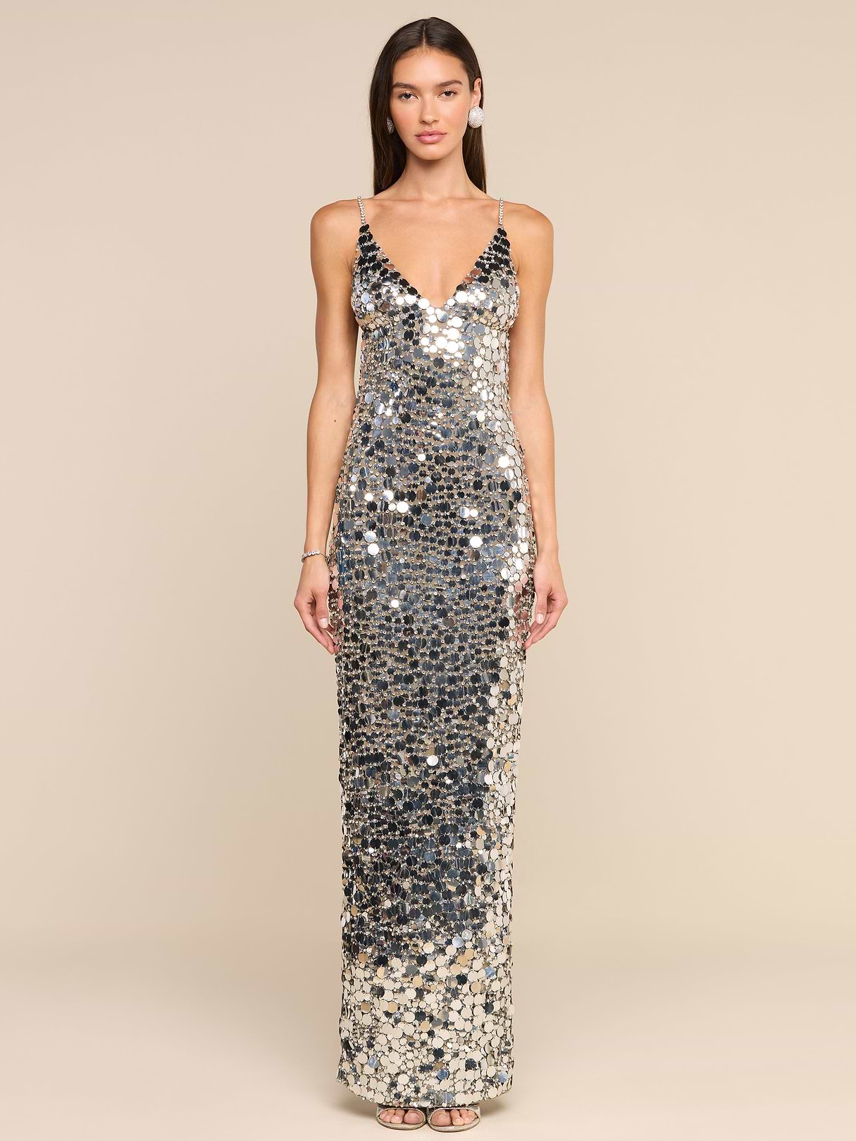 A woman stands facing forward, wearing the Caelith Sequin Gown—a sleeveless, floor-length silver sequined dress with a deep V neckline. She has straight, long brown hair and is posing against a plain white background.