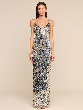 A woman stands against a beige background wearing the Caelith Sequin Gown—a sleeveless, floor-length dress adorned with large, reflective silver sequins, featuring a deep V-neckline and delicate spaghetti straps for a look inspired by timeless Hollywood glamour.