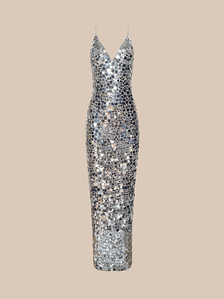 Channeling classic Hollywood glamour, the Caelith Sequin Gown features a sleeveless design with a deep V-neckline and delicate thin straps. Its silver sequined fabric hugs the body in a fitted silhouette, while the floor-length hem creates an effortlessly sophisticated look, beautifully displayed against a beige background.