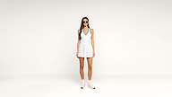A woman wearing sunglasses, a white tennis dress with a green collar, white socks, and sneakers stands against a plain white background.