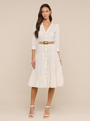 A woman wearing the Serafine Midi Dress—white, lightweight voile, button-down with three-quarter sleeves, a tiered skirt, tan belt—and white heels stands against a beige background.