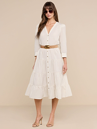 A woman stands confidently against a plain white background, wearing the Serafine Midi Dress in beige lightweight voile with a belt, paired with heeled sandals and sunglasses as she looks at the camera.