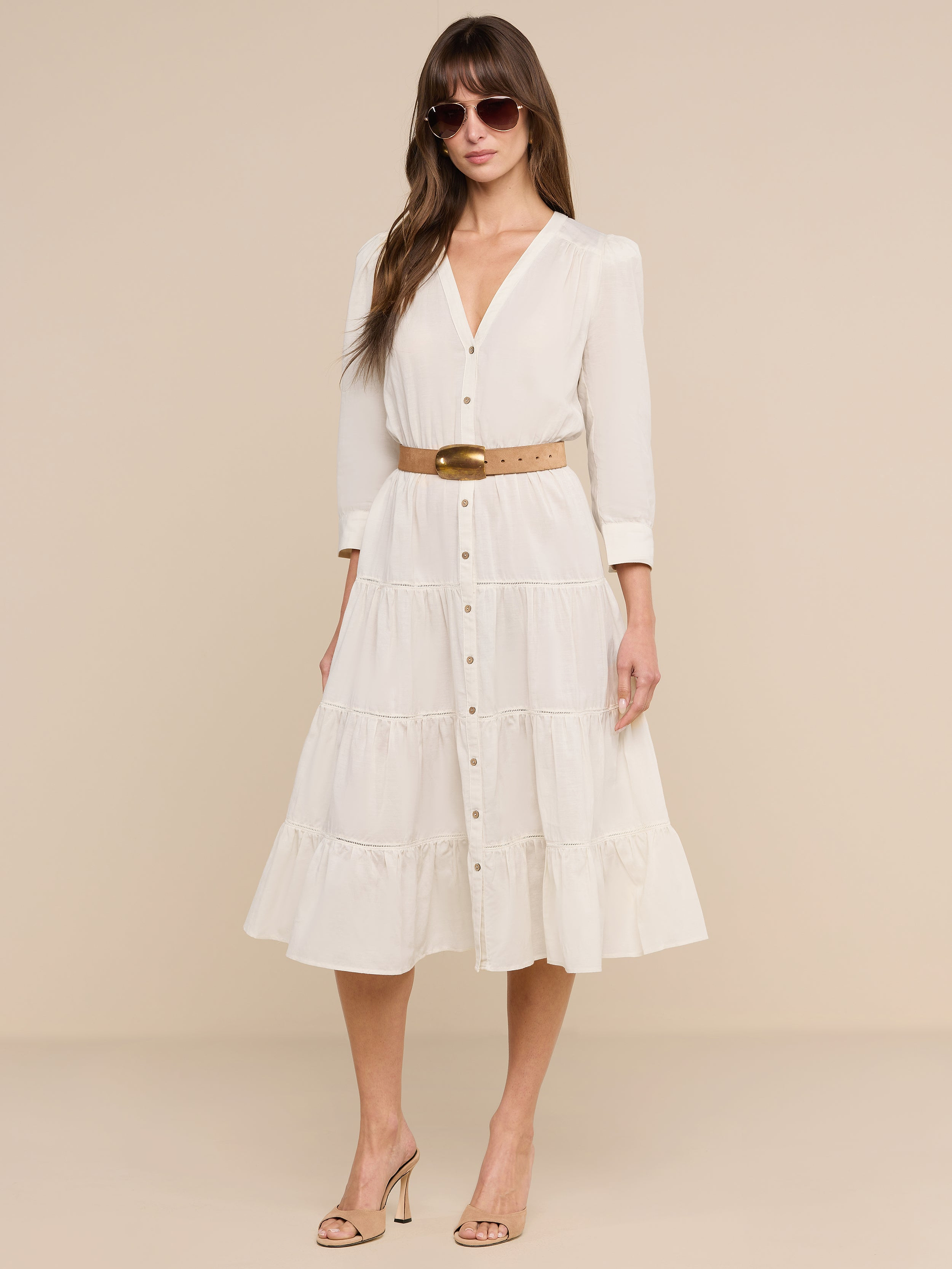 A woman stands confidently against a plain white background, wearing the Serafine Midi Dress in beige lightweight voile with a belt, paired with heeled sandals and sunglasses as she looks at the camera.