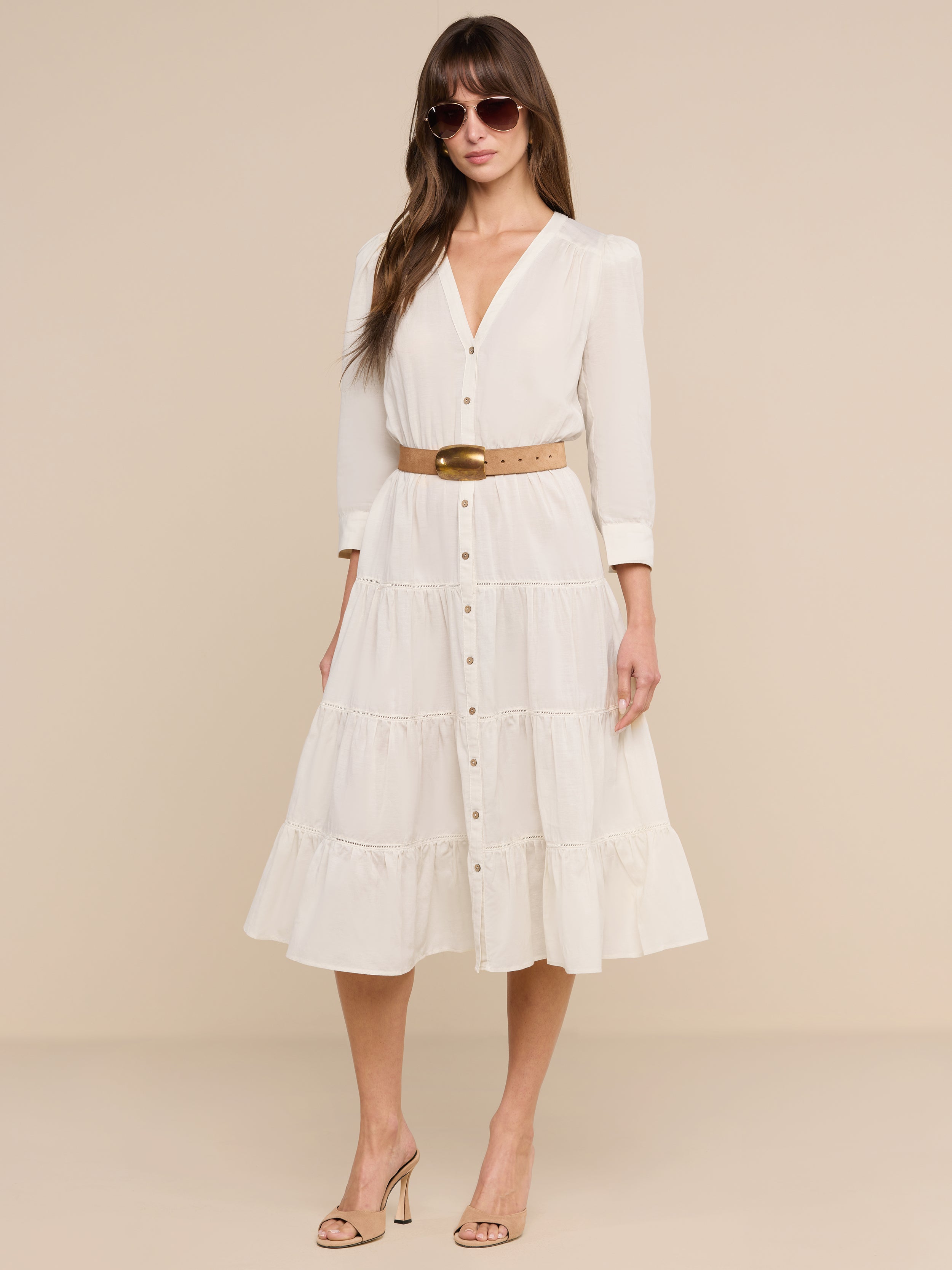 A woman stands confidently against a plain white background, wearing the Serafine Midi Dress in beige lightweight voile with a belt, paired with heeled sandals and sunglasses as she looks at the camera.