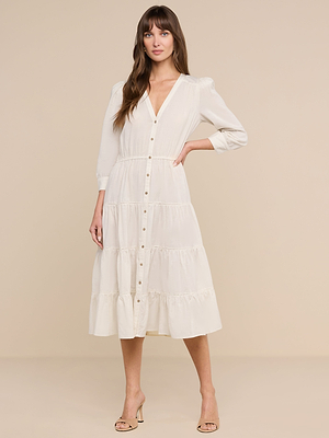 Una donna dai lunghi capelli castani posa su uno sfondo beige indossando labito Serafine Midi, caratterizzato da un modello bianco a maniche lunghe con gonna a balze. Completa il look con un paio di tacchi color carne.