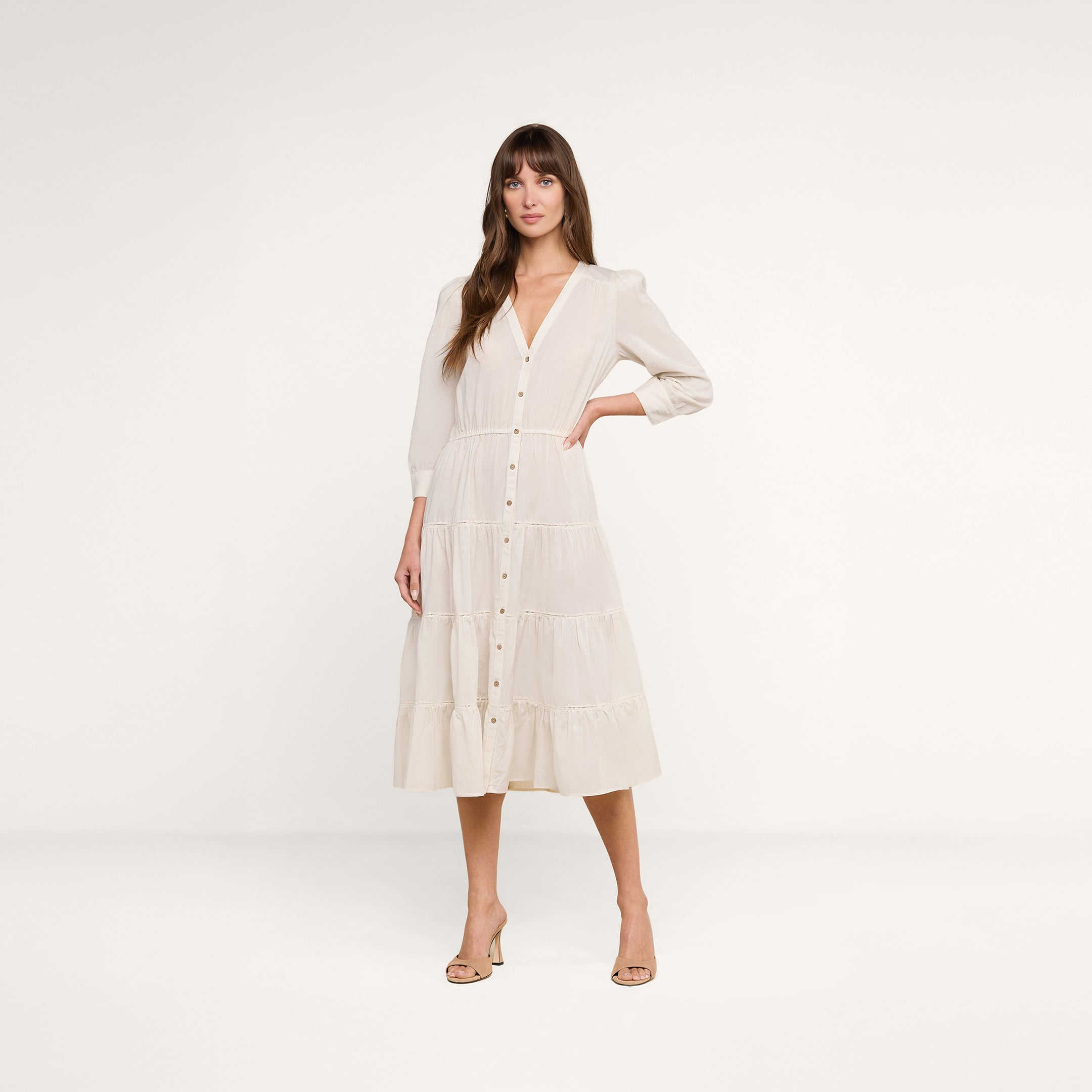 A woman stands on a white background wearing the Serafine Midi Dress—a cream-colored, lightweight voile dress with long sleeves, a button-down front, and a tiered skirt—paired with beige heeled sandals. She has long brown hair and poses with one hand on her hip.