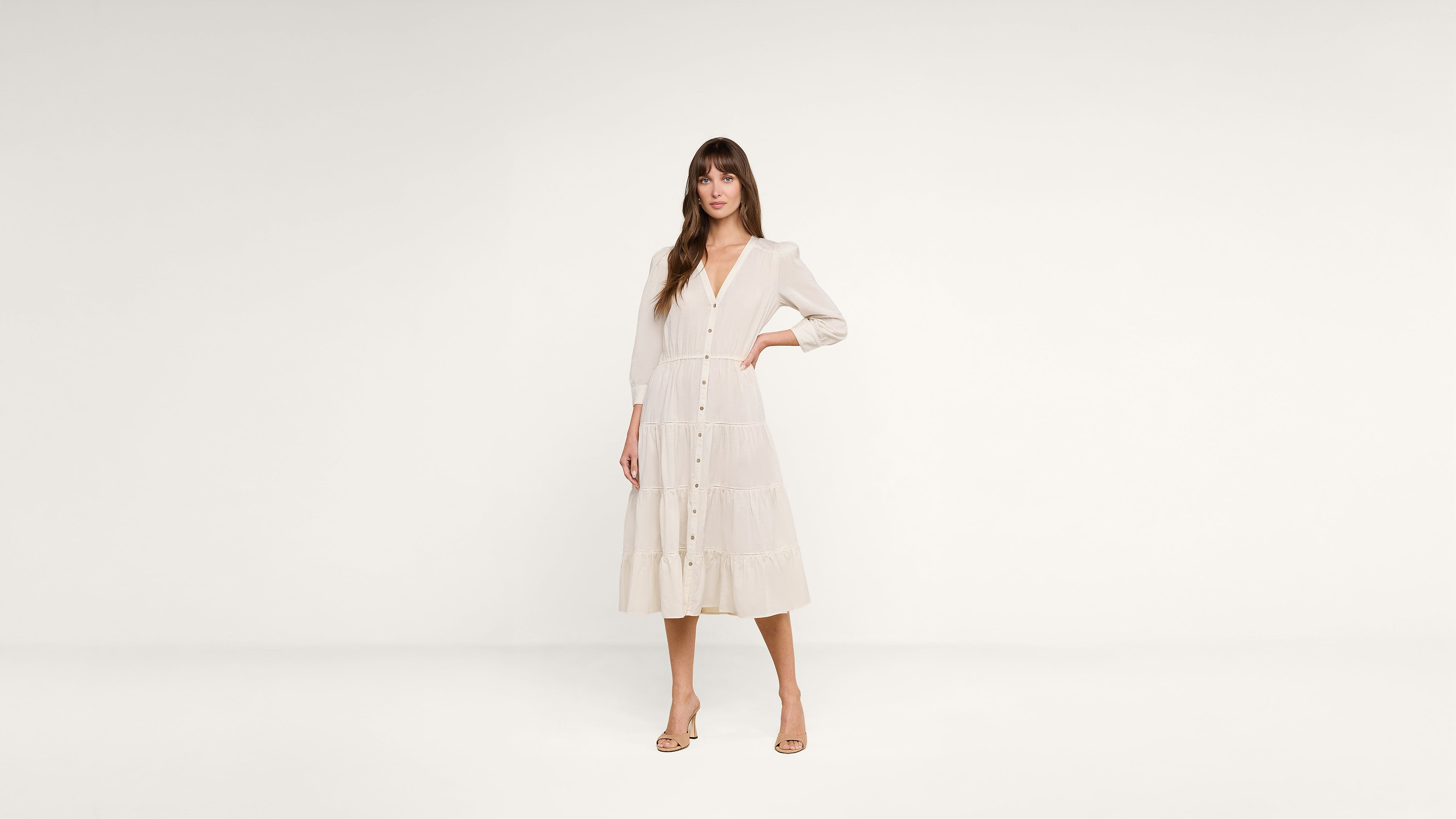 A woman stands against a plain white background wearing a long-sleeved, button-front, cream-colored midi dress and beige heels.