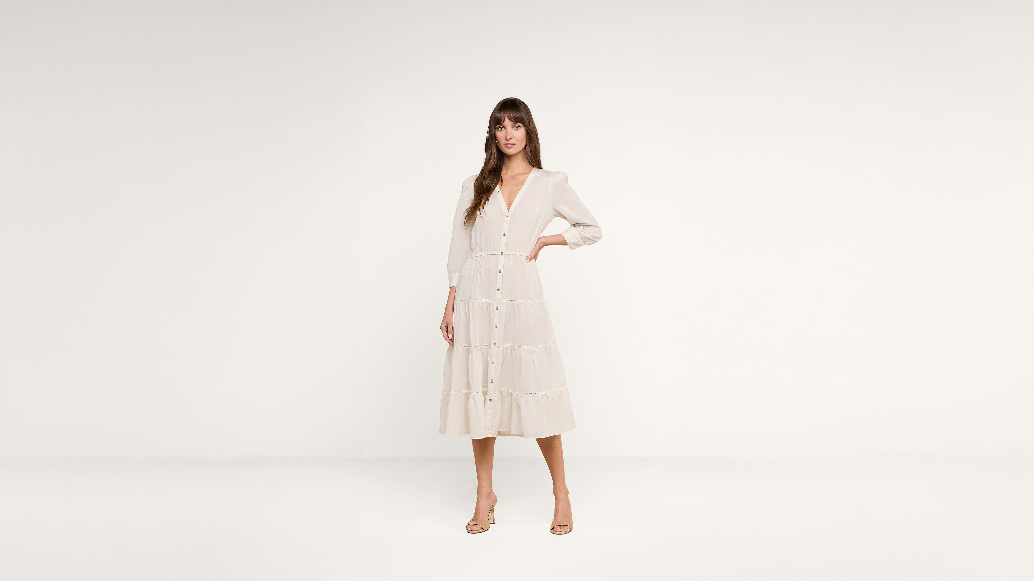 A woman stands against a plain white background wearing a long-sleeved, button-front, cream-colored midi dress and beige heels.