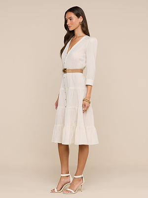 A woman stands in profile wearing the Serafine Midi Dress—a lightweight, cream voile summer dress with a tan belt, V-neck, three-quarter sleeves, and ruffled hem—styled with statement earrings, bracelets, and white heeled sandals.
