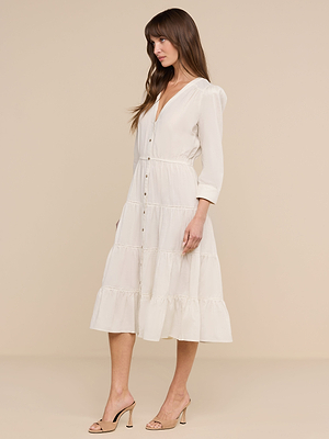 A woman stands sideways in a white Serafine Midi Dress with long sleeves, a v-neckline, and beige heels against a beige background.