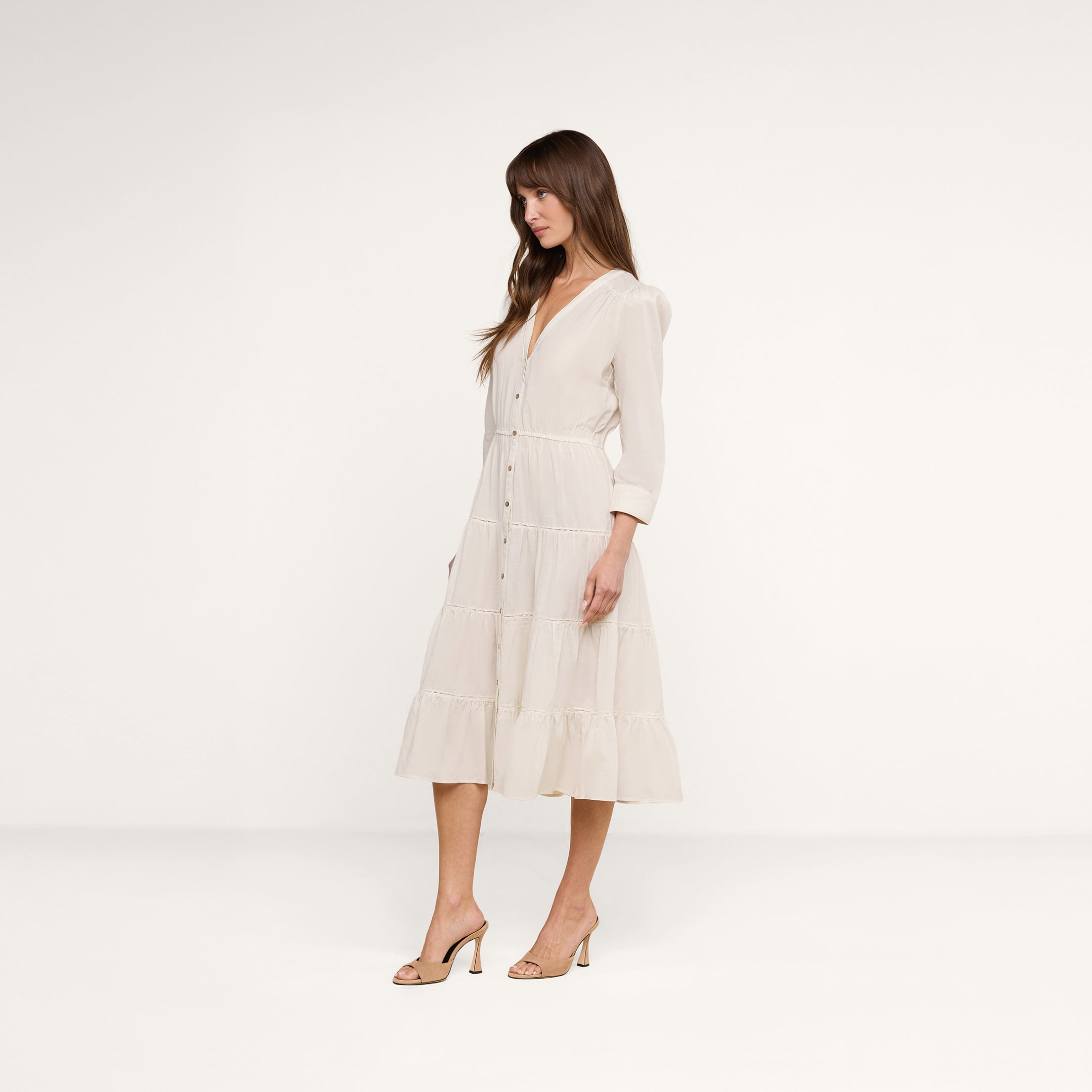 A woman stands sideways against a plain white background wearing the Serafine Midi Dress—a light-colored, long-sleeved, midi-length dress featuring a button-front detail and a tiered skirt—paired with heeled sandals.