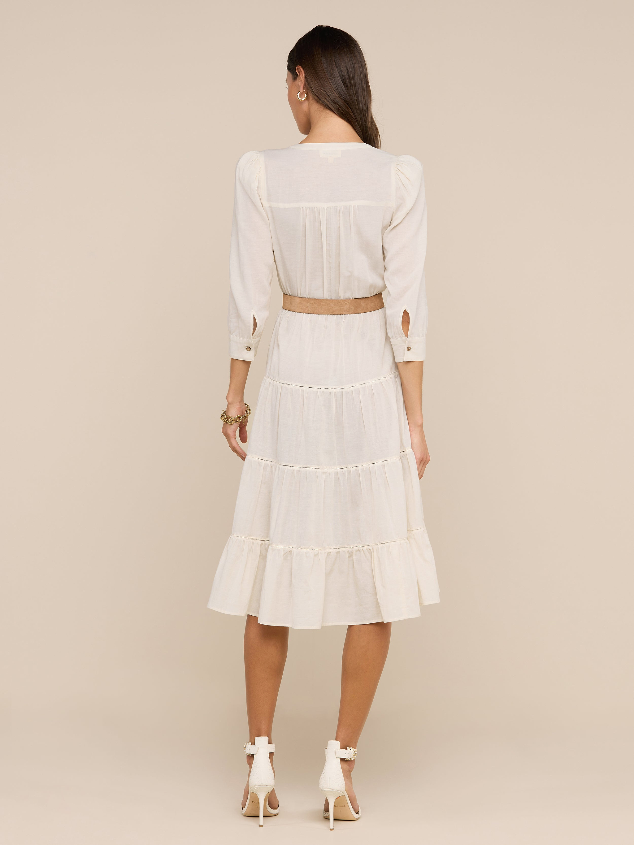 A woman faces away from the camera, modeling a light-colored Serafine Midi Dress with long sleeves and beige heels, set against a plain white background.