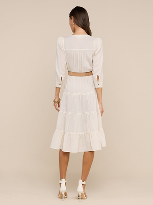 A woman faces away, wearing the white Serafine Midi Dress with long sleeves, tiered lightweight voile and ruffle details, paired with cream high-heeled sandals against a beige background.