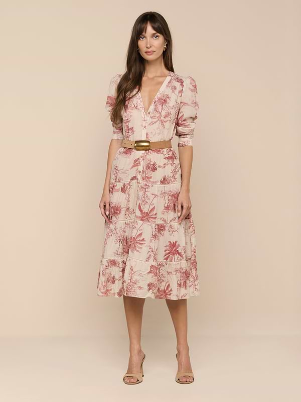 A woman with long brown hair stands against a plain white background wearing the Serafine Midi Dress, a light pink floral style featuring lightweight voile fabric, a V-neckline, puff sleeves, and a coordinating tan belt. She completes her look with nude heels.