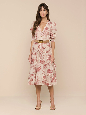 A woman with long brown hair and a neutral expression wears the Serafine Midi Dress—light pink with a floral pattern, puffed sleeves, v-neckline—styled with a brown belt and nude heels, standing against a beige background.