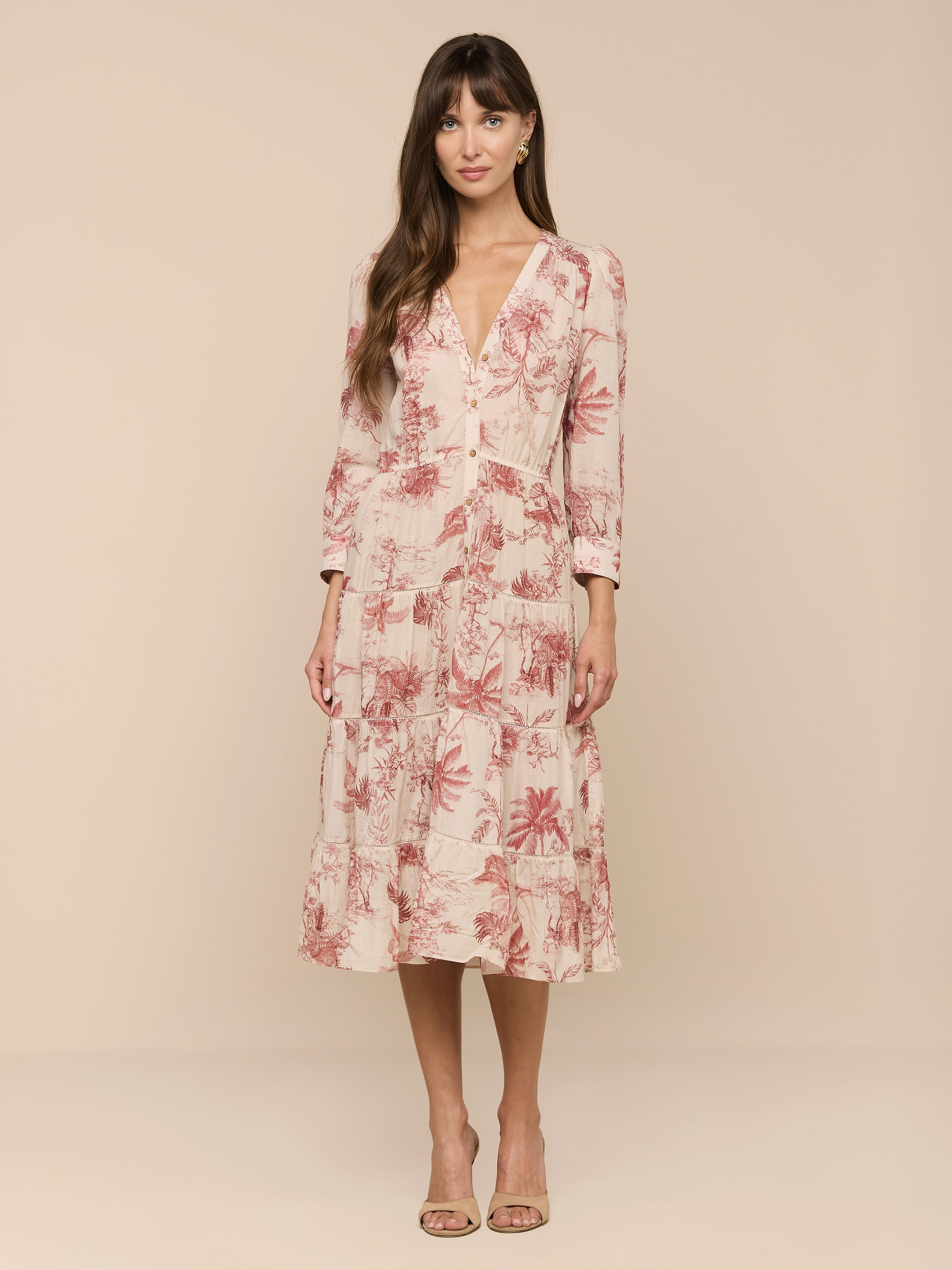 A woman stands against a plain white background wearing the Serafine Midi Dress—a light pink, long-sleeve midi dress with a red jungle print crafted from lightweight voile—paired with nude heels.