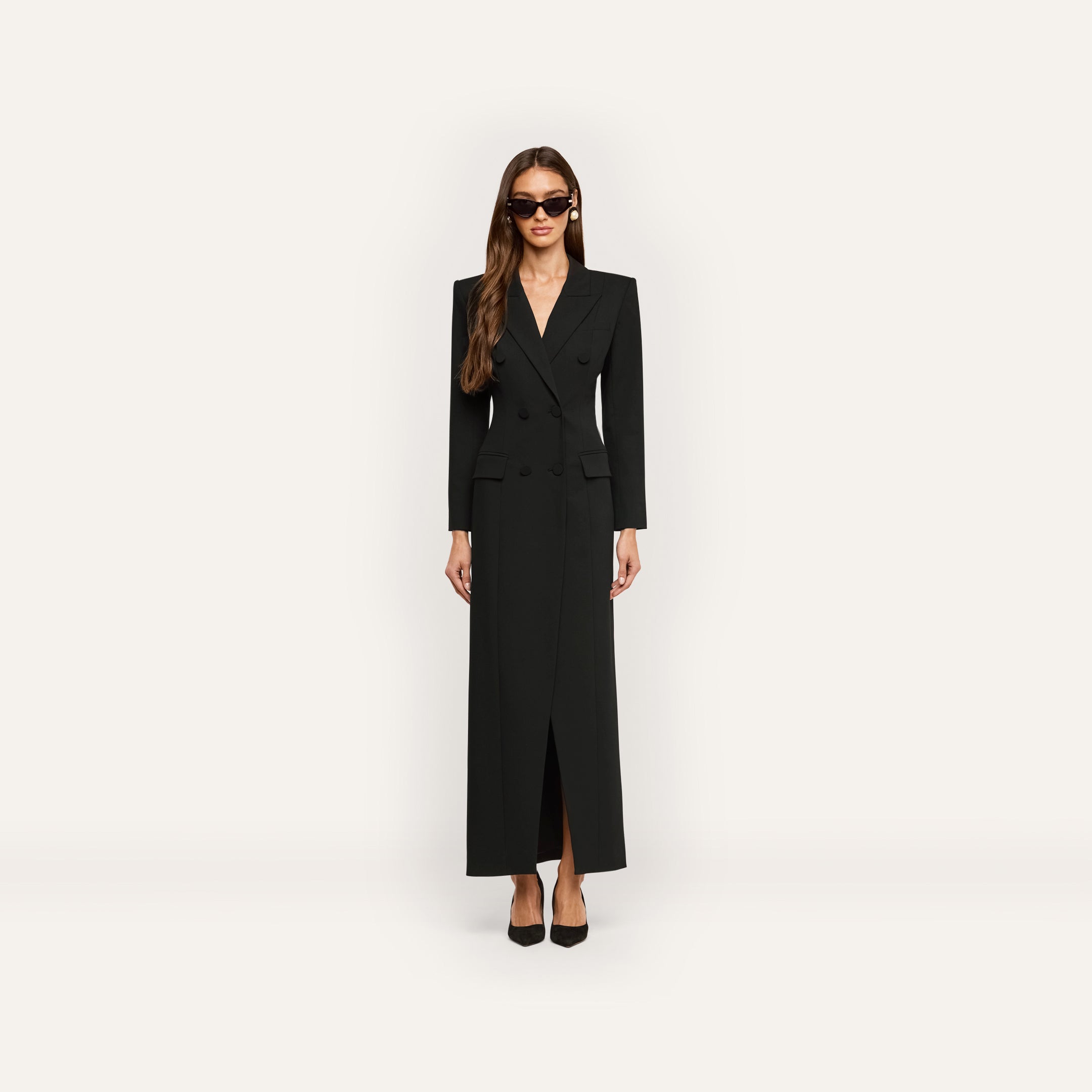 A woman wears a black, floor-length, long-sleeved Vale Tuxedo Dress in a double-breasted style, paired with black pointed-toe heels and sunglasses.