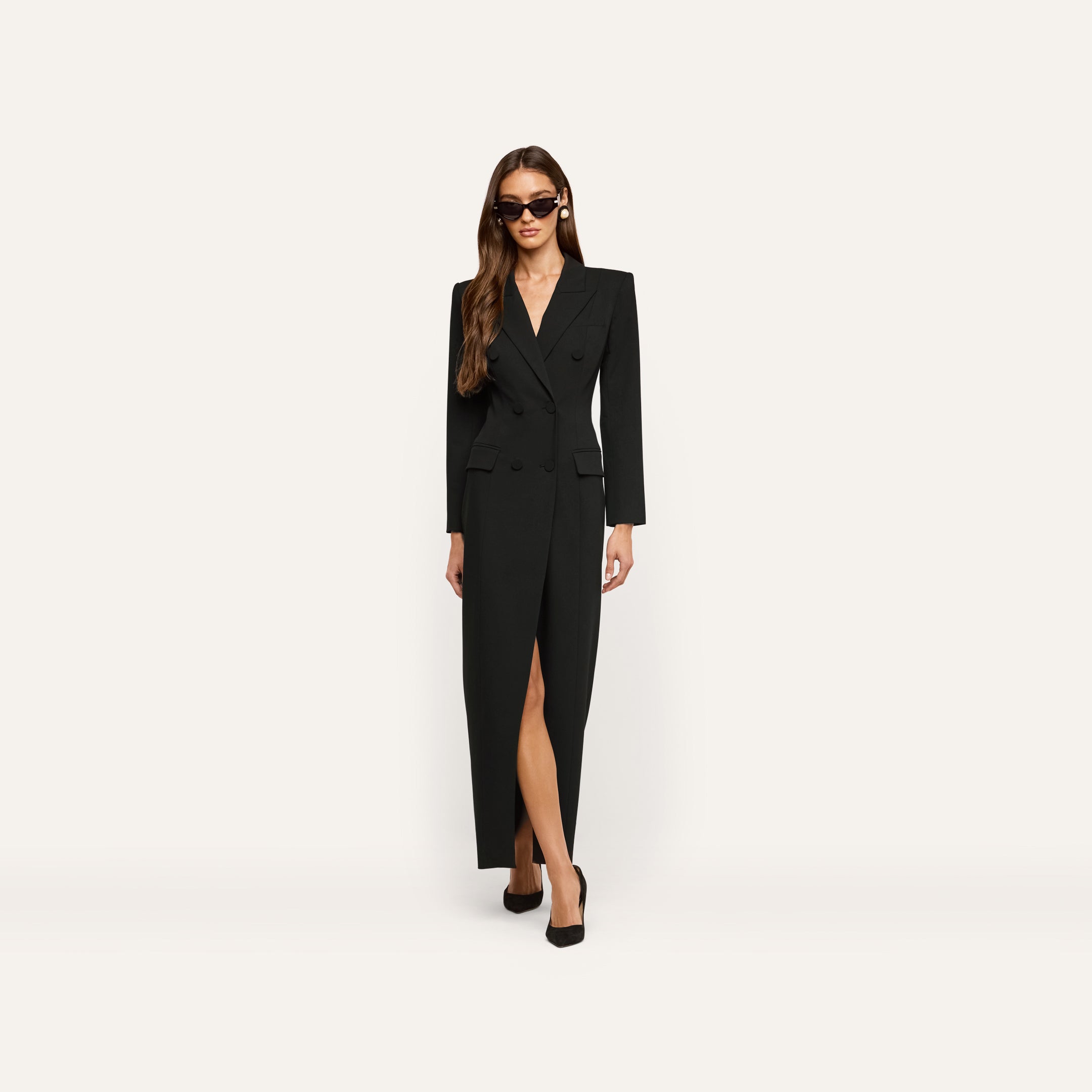 A woman wears a black, long-sleeved, double-breasted Vale Tuxedo Dress with a front slit and black pumps.