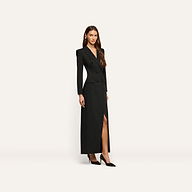 A woman wears the black, long-sleeved, double-breasted Vale Tuxedo Dress featuring a front slit and black pointed-toe heels, styled as a sophisticated maxi dress.