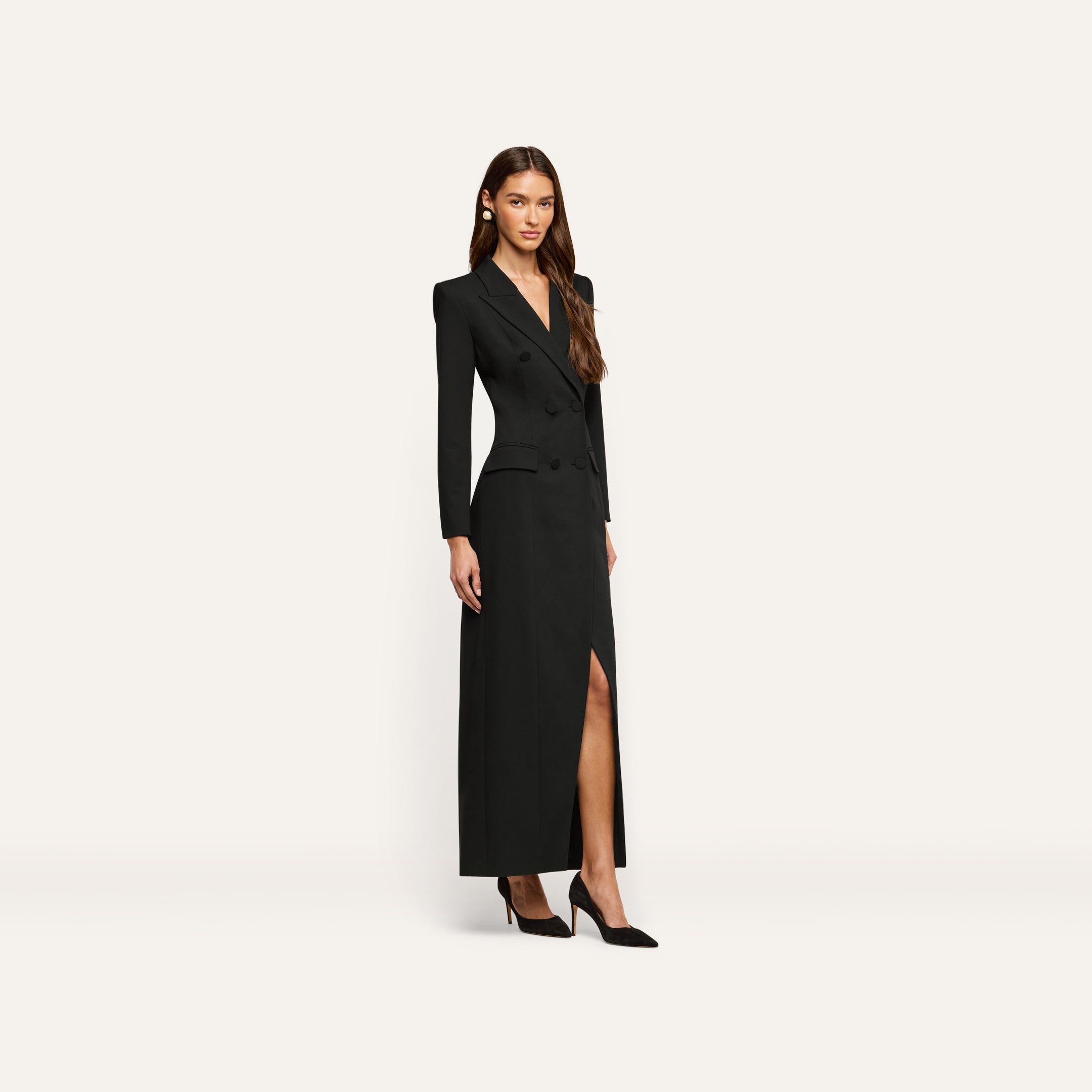 A woman wears the black, long-sleeved, double-breasted Vale Tuxedo Dress featuring a front slit and black pointed-toe heels, styled as a sophisticated maxi dress.