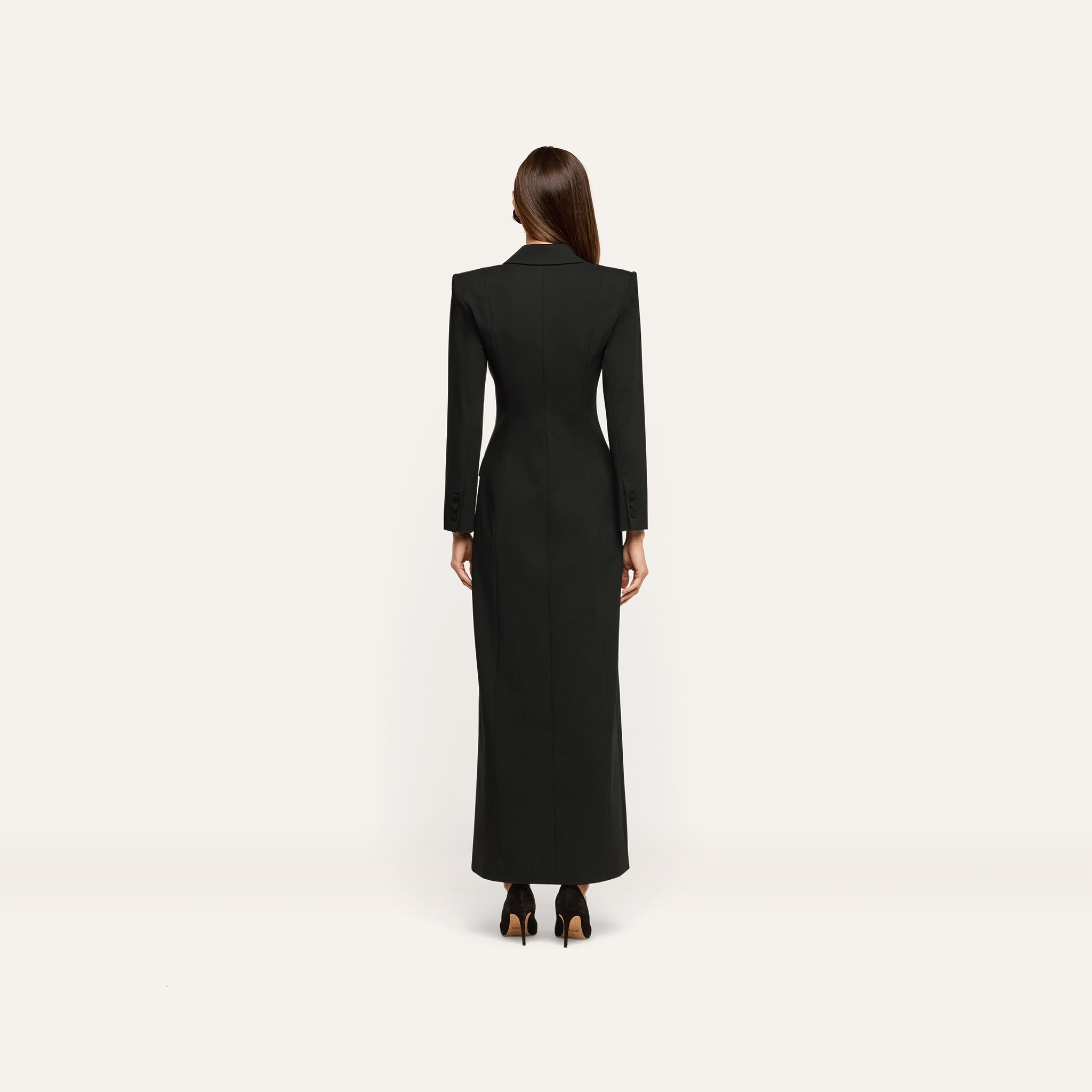 Back view of a person wearing a Vale Tuxedo Dress, a floor-length black maxi with structured shoulders and high heels.
