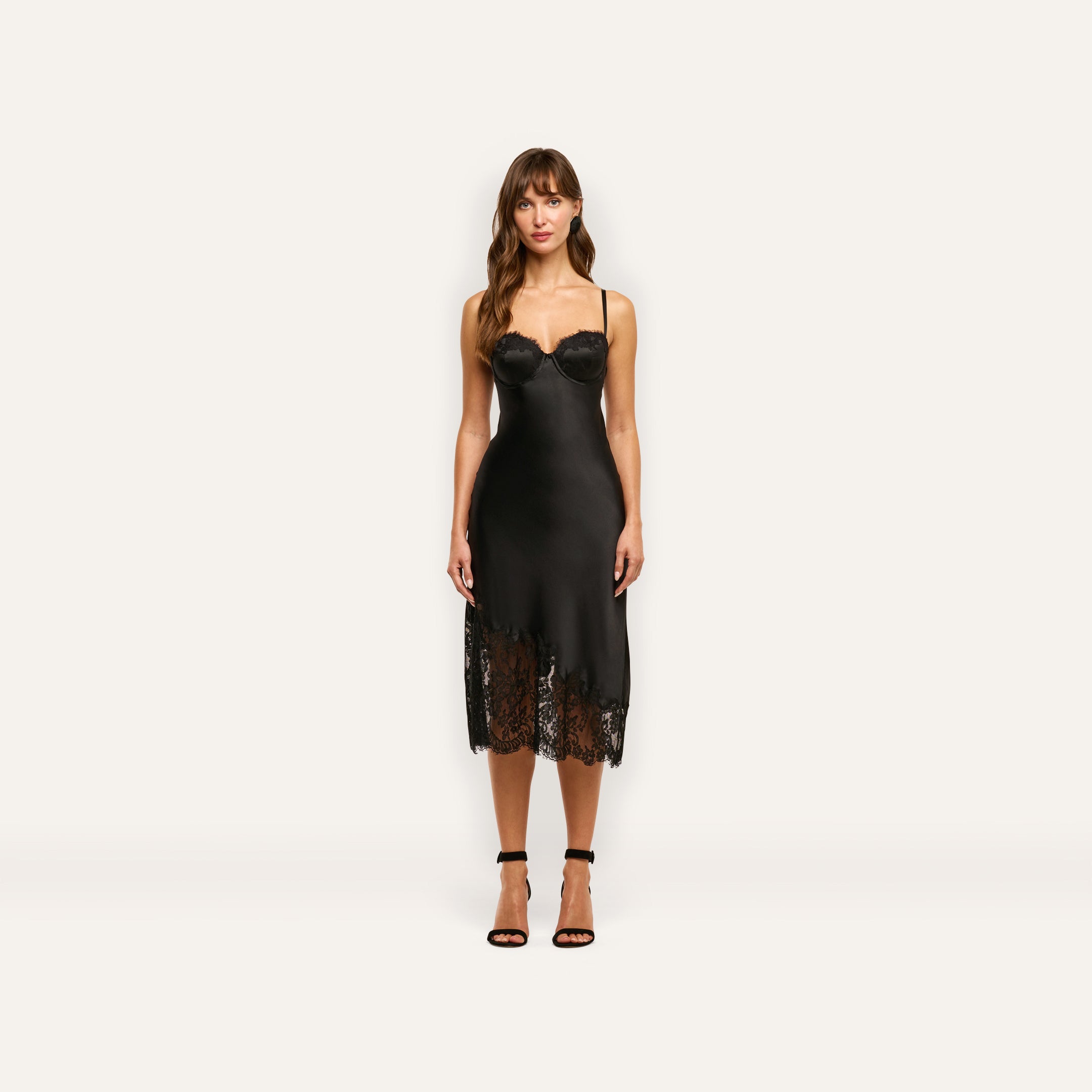 A woman wears the Adrienne Silk Lace Dress, a black silk midi-length slip dress with a corset-style bodice and lace trim at the hem, paired with black ankle-strap heels.
