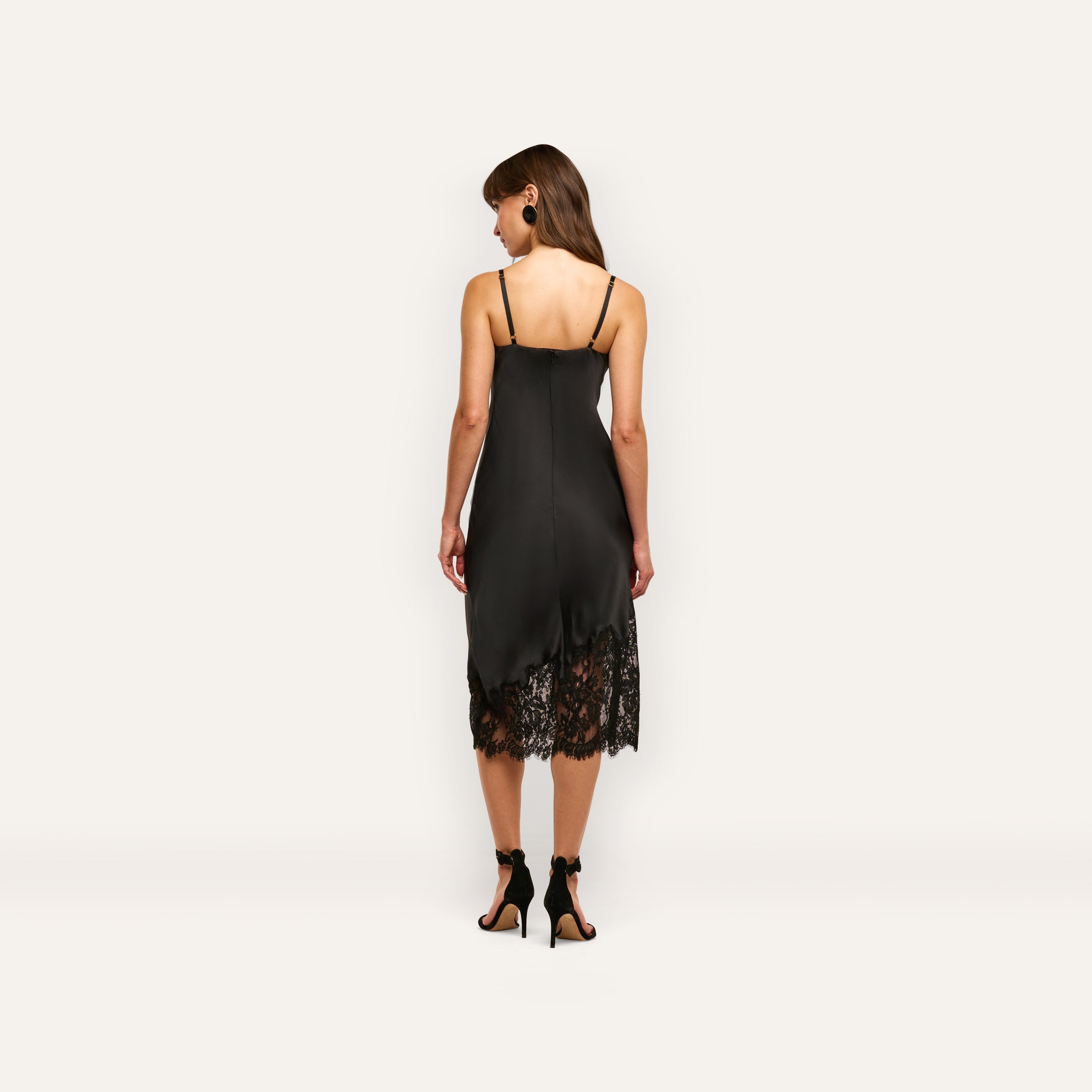 Back view of a woman wearing the black Adrienne Silk Lace Dress, a midi-length slip dress with spaghetti straps and a black lace hem, paired with black high-heeled sandals.