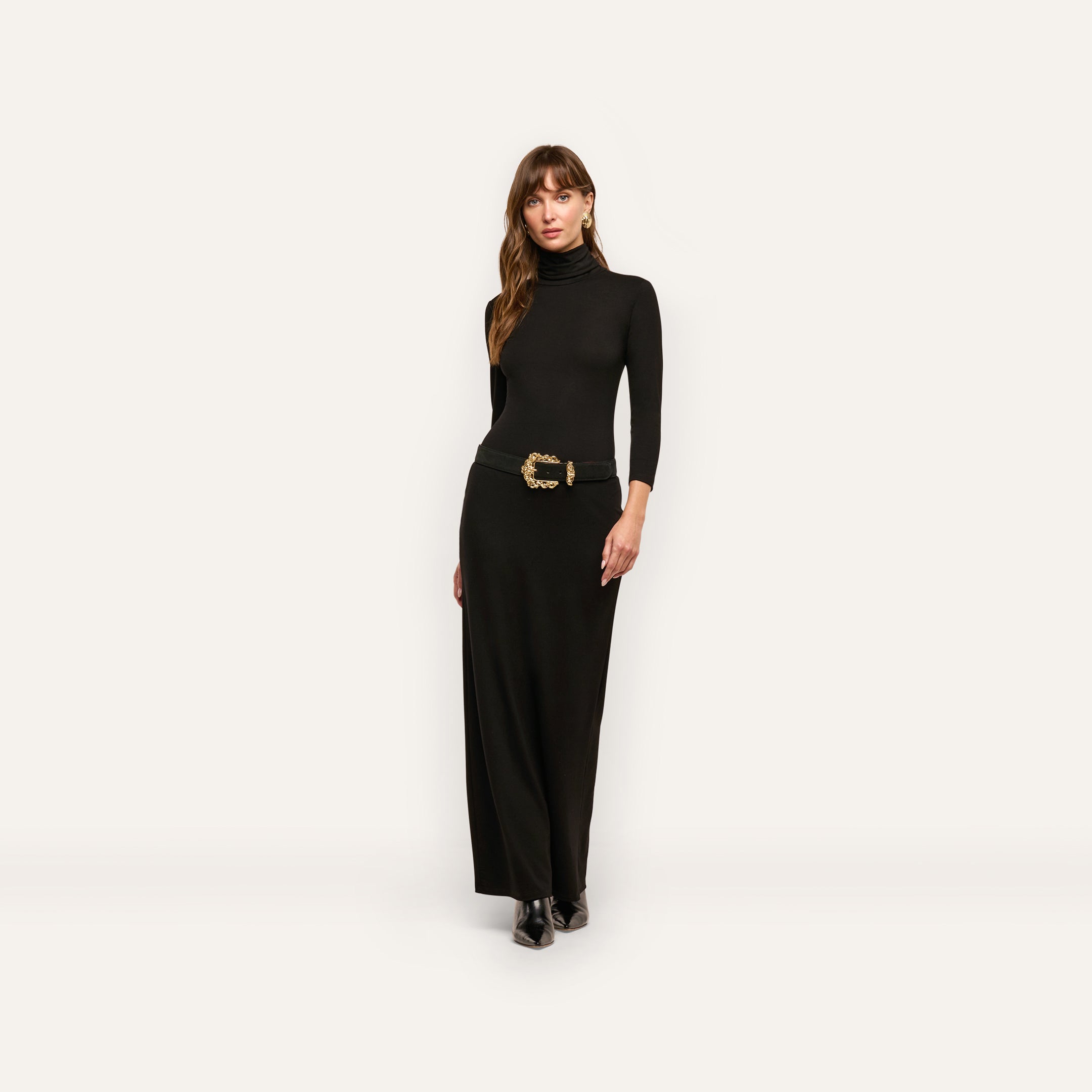 A woman wears a long-sleeved black jersey knit Jaesyn Dress turtleneck maxi dress with a black belt featuring a decorative gold buckle.