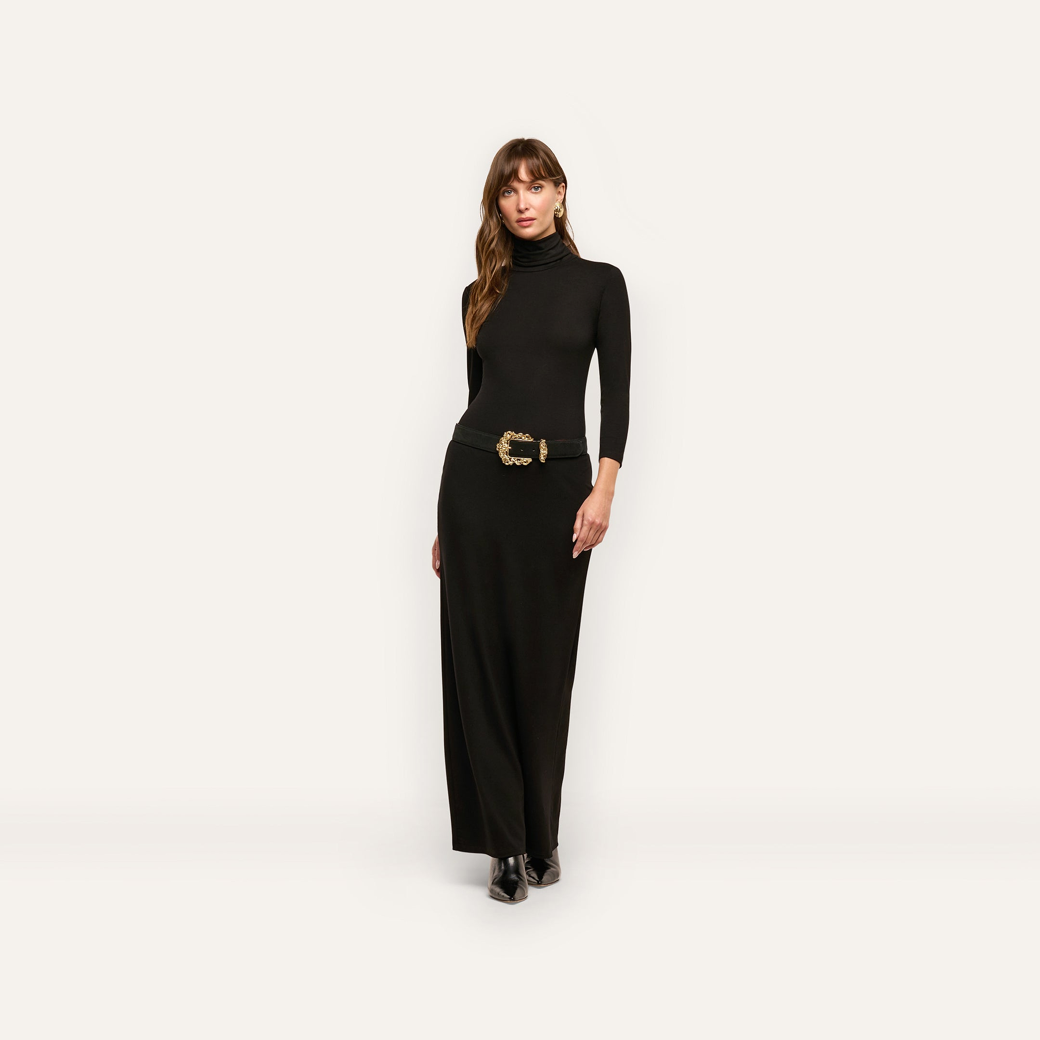 A woman wears a long-sleeved black jersey knit Jaesyn Dress turtleneck maxi dress with a black belt featuring a decorative gold buckle.