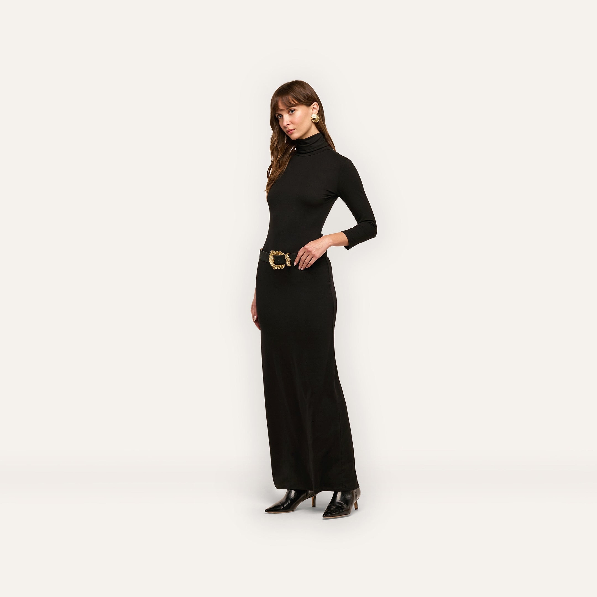 A woman wears the Jaesyn Dress, a black jersey knit turtleneck maxi dress with three-quarter length sleeves and a wide belt featuring a decorative gold buckle.