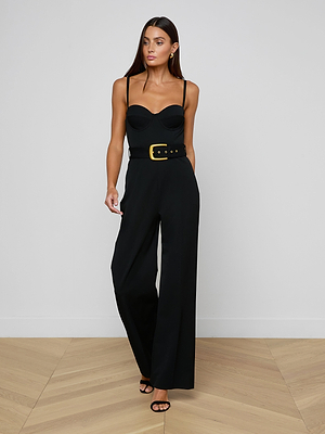 A woman stands on a wooden floor against a plain white wall, wearing the Jessa Bustier Jumpsuit—black bi-stretch twill with wide legs and a waist-defining belt featuring a gold buckle.