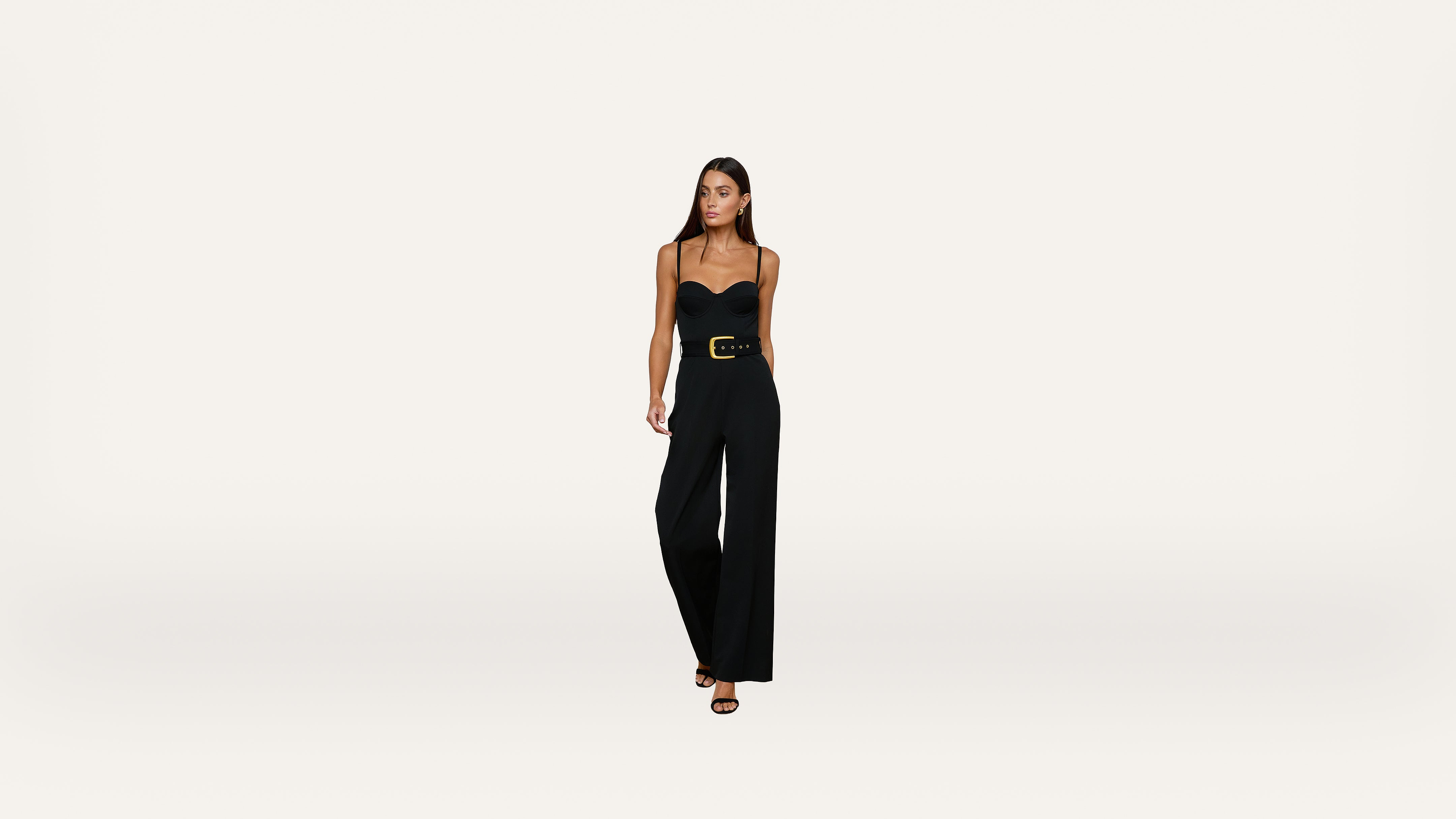 A woman wearing a strapless black jumpsuit with a wide belt and black heels stands against a plain white background.