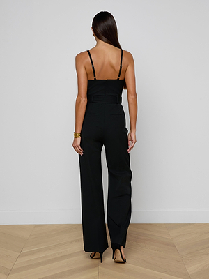 A woman with long, straight hair faces away, wearing the sleek black Jessa Bustier Jumpsuit and black heels. She accessorizes with gold bracelets and stands on a light wood herringbone floor against a plain white wall.
