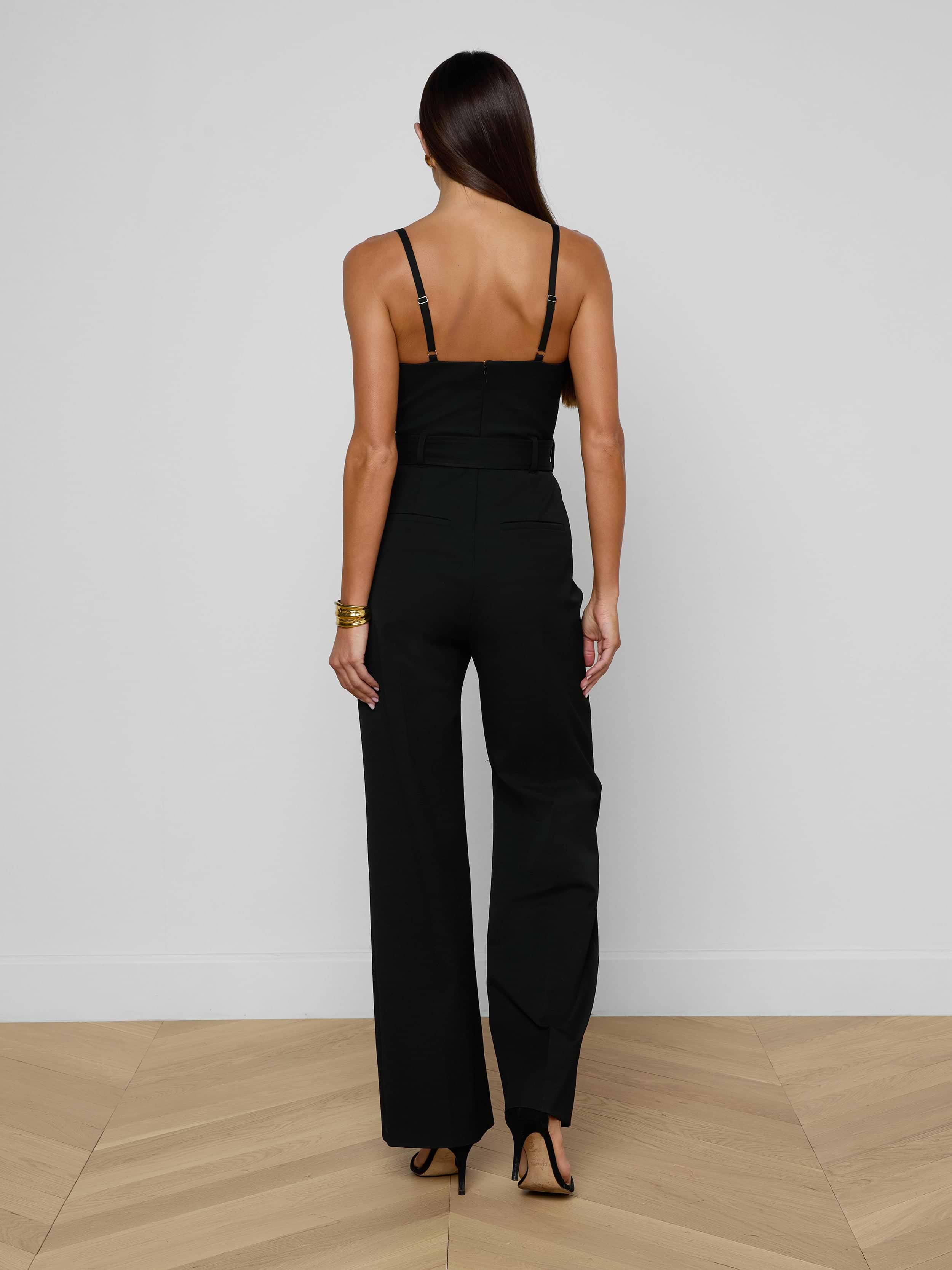 A woman stands with her back to the camera, wearing the Jessa Bustier Jumpsuit in black bi-stretch twill, paired with black heels, set against a plain light background.