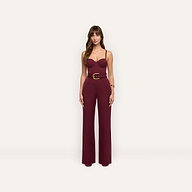 A woman wears a burgundy, sleeveless, wide-leg Jessa Bustier Jumpsuit featuring a corset-style bodice and a gold-buckled belt.