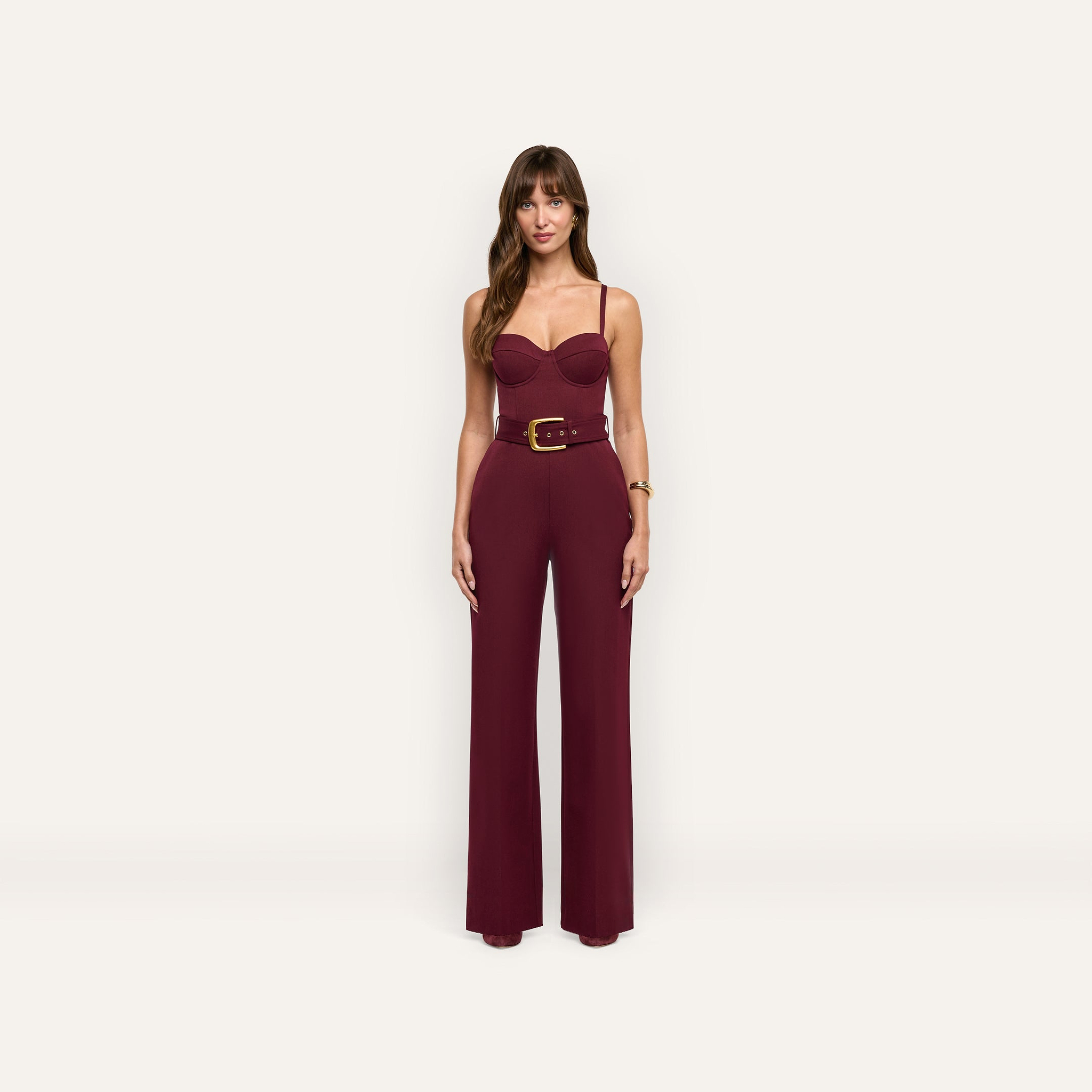 A woman wears a burgundy, sleeveless, wide-leg Jessa Bustier Jumpsuit featuring a corset-style bodice and a gold-buckled belt.