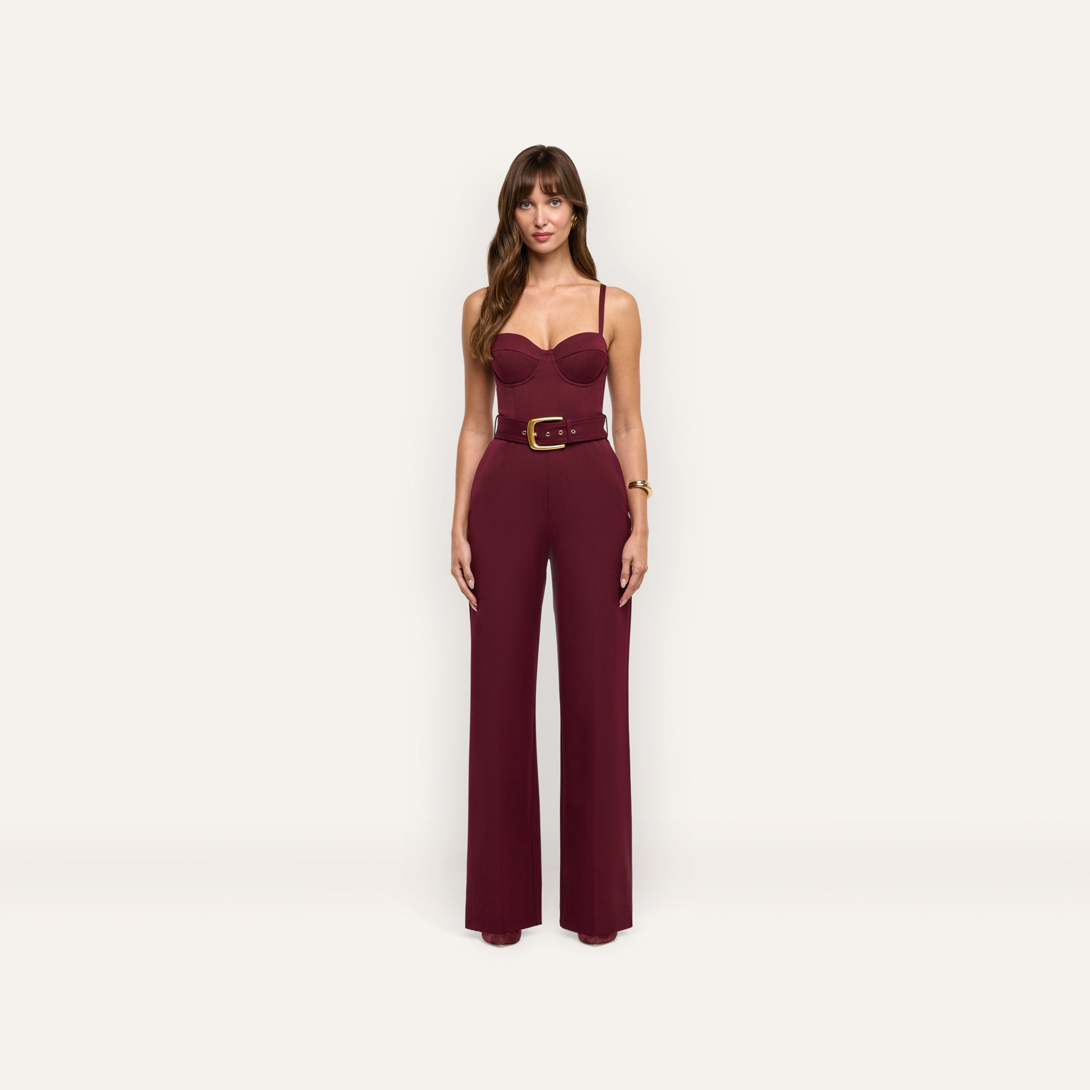 A woman wears a burgundy, sleeveless, wide-leg Jessa Bustier Jumpsuit featuring a corset-style bodice and a gold-buckled belt.