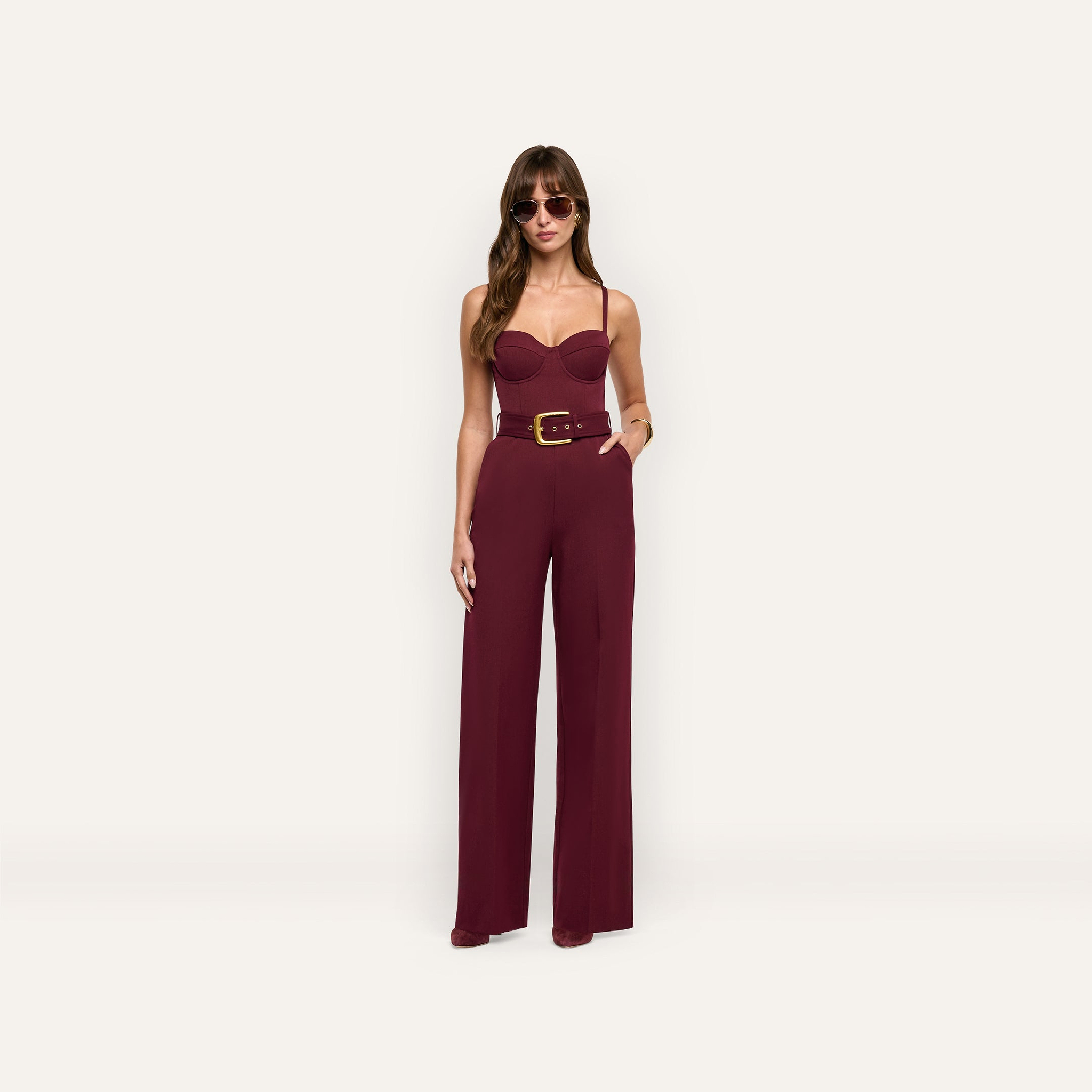 A woman wearing a burgundy Jessa Bustier Jumpsuit with a belted waist and wide-leg trousers, styled with sunglasses.